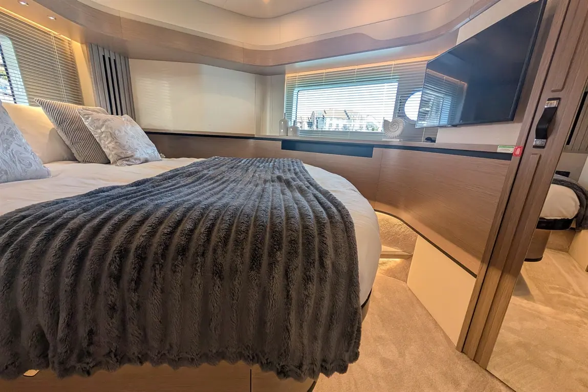 Stylish bedroom with a large bed, grey throw, decorative pillows, and a TV, featuring soft lighting and a window view.